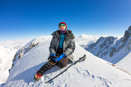 woman climber in helmet and down jacket with trekking sticks sits on top of a mountain.の写真素材