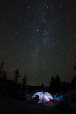White tent under the starry sky and the milky way in the trees in the winter mountains at night.