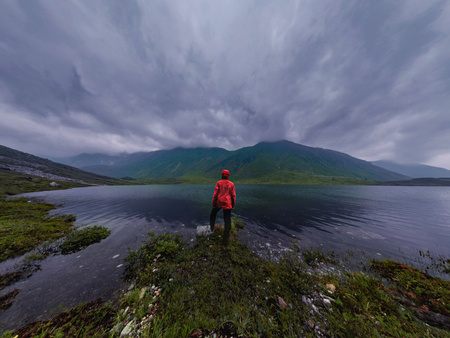 man stands by lake in red jacket in cloudy weather panorama