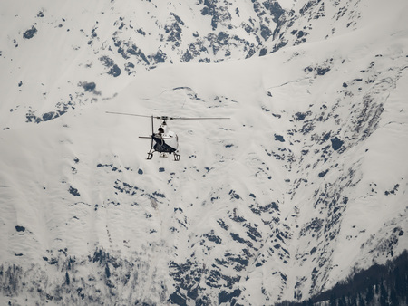 White helicopter flies over snowy mountains on a cloudy day