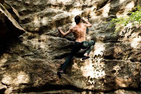 Rock climber climbs bouldering on a cliff on forest. Low angle of strong rock climbing man hanging free on rock with sunflareの写真素材