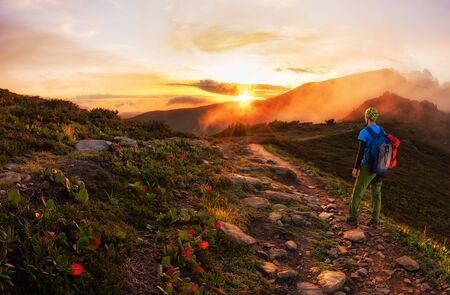 Hiker with backpack walking along a mountain trail in the background of dawn.