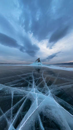 Ice of Lake Baikal in cracks near the island of Elenka at sunset under gray clouds. Wide panoramaの写真素材