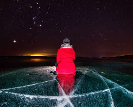 girl in red sits on an ice-lit frozen Lake Baikal under a starry night sky with the constellation...