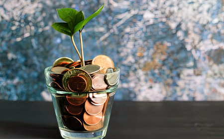 Plant growing out of coins in a glass vase on a blue backgroundの素材
