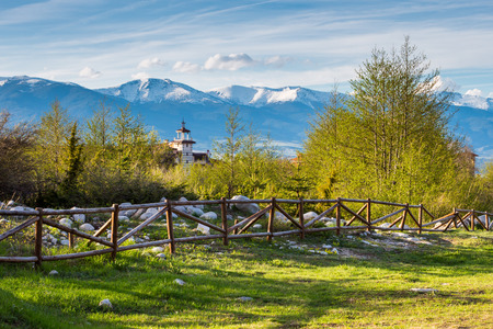 Spring landscape with wooden fence, trees, part and snowy mountainsの写真素材