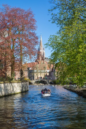 Bruges, Belgium - April 10, 2016: Canal and bridge view, boat with tourists, colorful spring trees and church tower in popular belgian destinationのeditorial素材