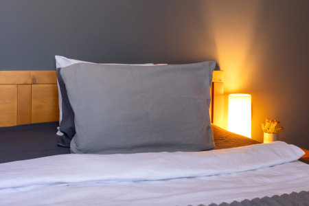 Cozy bedroom interior with grey pillows, white linens, wooden headboard and warm bedside lamp...
