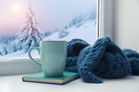 Winter background - cup with candy cane, woolen scarf and gloves on windowsill and winter scene outdoors. Still life with concept of spending winter time at cozy home with cold weather outdoorsの写真素材