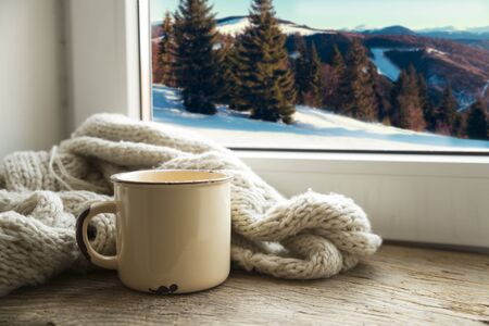 Winter background - cup with candy cane, woolen scarf and gloves on windowsill and winter scene outdoors. Still life with concept of spending winter time at cozy home with cold weather outdoorsの写真素材