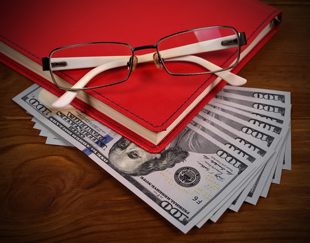 red leather diary, glasses and dollar bills on wooden deskの写真素材