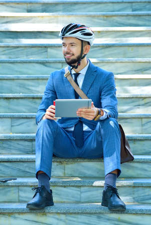 Businessman in helmet sitting on stairs with cup of coffee and using digital tablet. Business and alternative transport conceptの写真素材