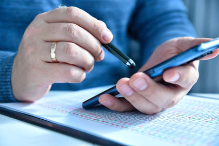 Close up of trader checks financial report and compares data on smartphone and paper. Internet...
