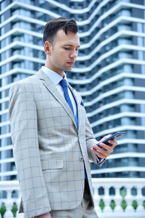 Young businessman in suit using mobile phone on a modern skyscraper background. Business and technology concept. Close upの写真素材