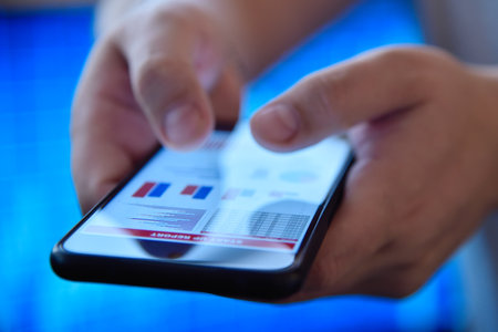 Hands using mobile phone displaying chart financials, against a vibrant blue stock market background. Concept of economy and trading. Close upの写真素材