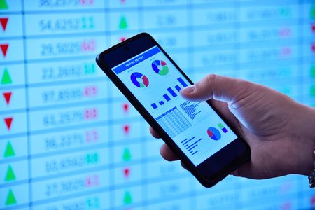 Businessman hand using a smartphone displaying financial charts, against a vibrant blue stock market background. Concept of tradingの写真素材