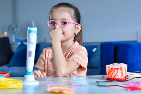 Little girl in pink glasses sits at table and prepares to make craft with 3d pen. Concept of creativity and innovation in art.の写真素材