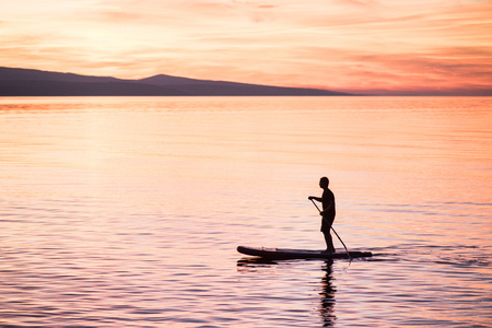 Silhouette of man at sunset standing on paddle board. Summer beach leisure activity. Active healthy lifestyle and travel summer conceptの写真素材