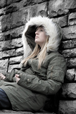 young woman sat against a stone wall looking up with a hood onの写真素材