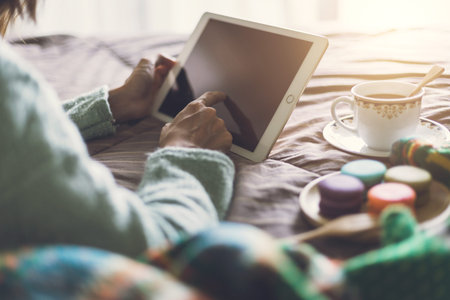 Young woman using tablet at cozy home atmosphere on the bed with cup of coffee and macaroon. Soft focusの写真素材