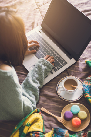 Young woman using laptop at cozy home atmosphere on the bed with cup of coffee and macaroon.の写真素材