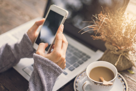 Young woman working by using cellphone and laptop on wooden table with coffee and flowerの写真素材