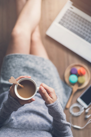 Young woman relaxing at cozy home atmosphere on the wooden floor with cup of coffee and macaroon.の写真素材