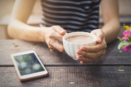 Female hands holding a cup of coffee with foam on wooden table in the gardenの写真素材
