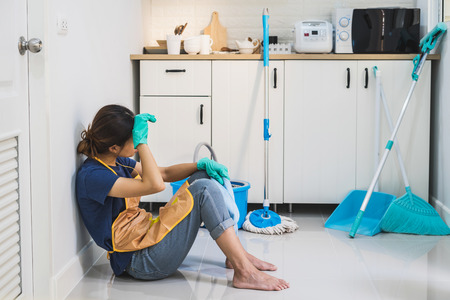 Tired young woman sitting on kitchen floor with cleaning products and equipment, Housework concept