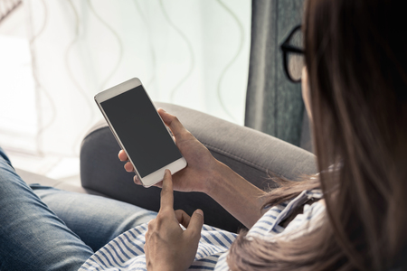 Young woman in glasses siiting on sofa and using smart phone at homeの写真素材