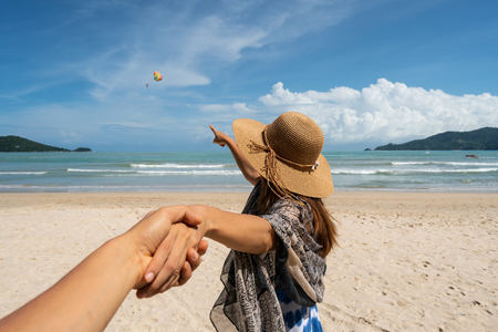 Young woman traveler holding man's hand and looking parachute flying in the sky on the beach, Couple vacation travel summer conceptの写真素材