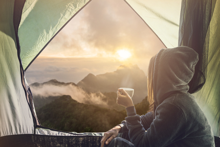 Young woman traveler sitting in the tent, relaxing and drinking coffee with beautiful sunriseの写真素材