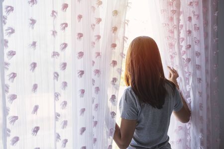 Young asian woman unveil curtain and looking out of window in the morningの写真素材