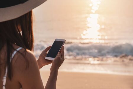 Young woman traveler using mobile phone on tropical beach at sunset, vacation and summer conceptの写真素材