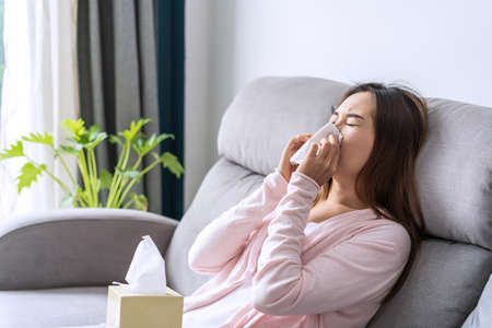 Young asian women with allergies feeling unwell using tissues and sneezing at homeの写真素材