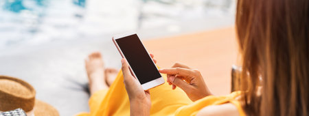 Young woman traveler relaxing and using a mobile phone by a hotel pool while traveling for summer...