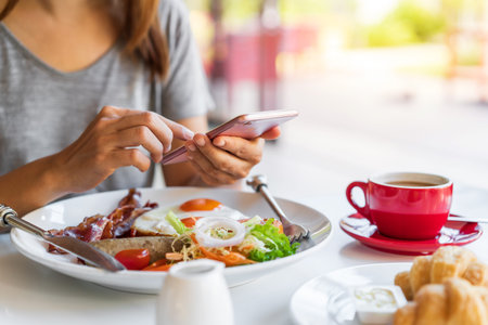 Young woman using smart phone with cup of coffee while eating breakfast