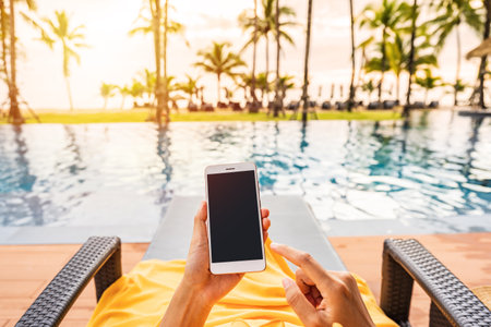 Young woman traveler relaxing and using a mobile phone by a hotel pool while traveling for summer vacationの写真素材