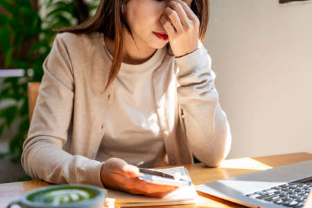Tired and stressed young Asian woman sitting at her working place with a cup of coffee and breakfast in the morningの写真素材