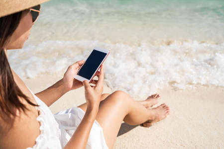 Young woman using mobile phone at beautiful tropical white sand beach with wave foam and transparent sea, Summer vacation and Travel concept with copy spaceの写真素材