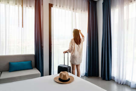 Young woman traveler with luggage looking at view in hotel room on summer vacationの写真素材