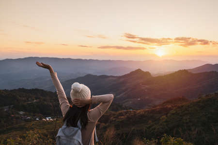 Happy young woman traveler relaxing and looking at the beautiful sunset on the top of mountainの写真素材