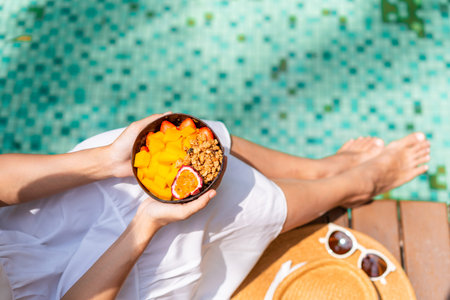 Young woman traveller relaxing and eating refresh bowl with fruits at tropical resort pool on summer...