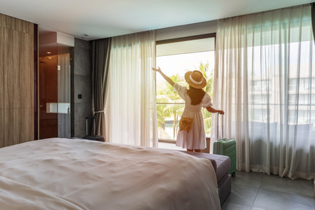 Young woman traveler opening the curtains and looking at the view from the window of a hotel room while on summer vacation, Travel lifestyle conceptの写真素材