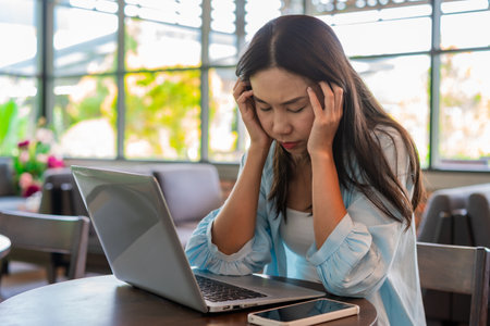 Tired and stressed young Asian woman working with laptop sitting at working spaceの写真素材