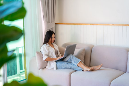 Young Asian female relaxing and using laptop on a sofa at homeの写真素材