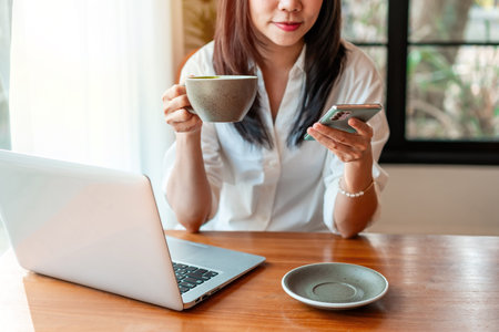 Smiling young Asian business woman using phone and working with laptop in cafe, Digital nomad lifestyleの写真素材