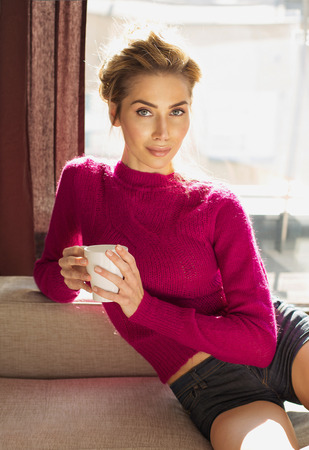 Beautiful blond woman sitting on coach in home and holding a cup の写真素材