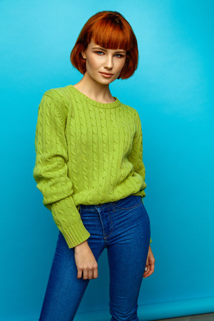 Colorful portrait of young female model with red hair and green sweater isolated on blue backgroundの写真素材