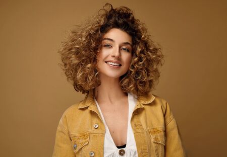 Happy young woman with curls posing at studio on brown studio background. Blonde woman with afro hairstyle smilingの写真素材